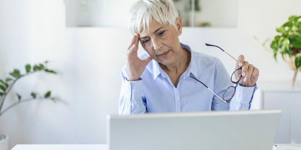 Senior frustrated woman working from home office in front of laptop, having a headache. Mature woman having a painful face expression