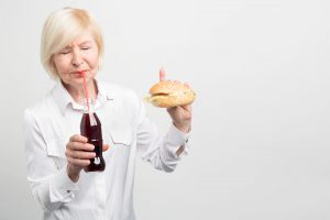 A picture of old woman tasting coca cola. and eating a burger. She doesn't like to have a healthy lifestyle. She prefers to eat tasty but fatning and bad food. Isolated on white background
