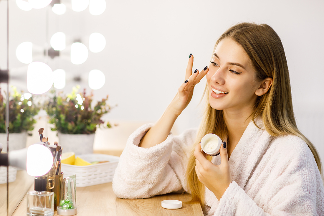 Young beautiful blonde girl cares for face skin with moisturizer in front of mirror.