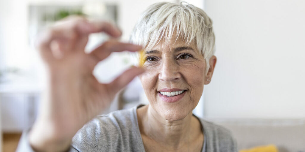 Senior woman at home sitting at table and kitchen at home holding cup of water and pill coronavirus treatment close-up blurred background