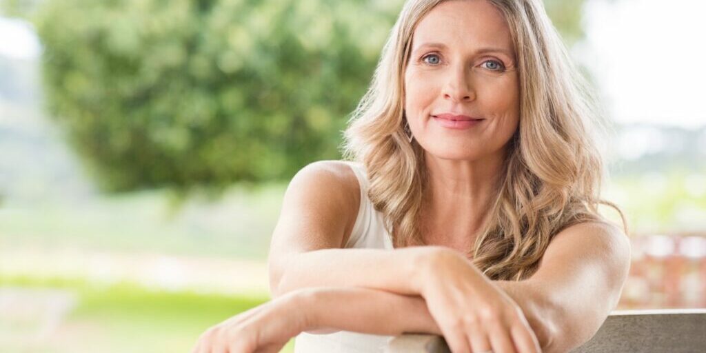Happy senior woman relaxing on bench in the lawn. Close up face of a mature blonde woman smiling and looking at camera. Retired woman in casuals sitting outdoor in a summer day.