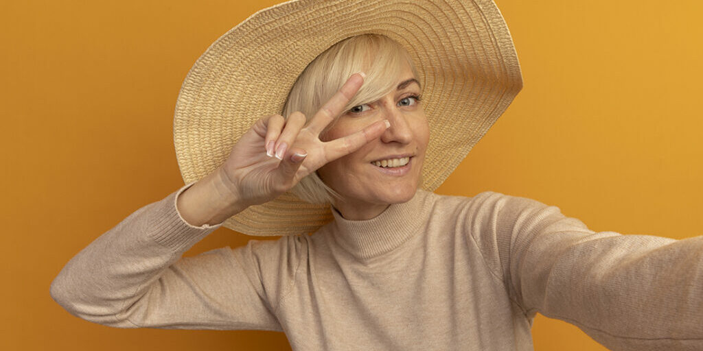 smiling pretty blonde slavic woman with beach hat gestures victory hand sign pretending to hold camera isolated on orange background with copy space