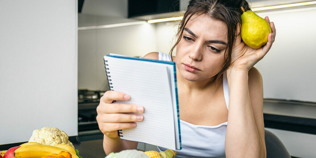 A young woman in the kitchen with a notebook among vegetables.
