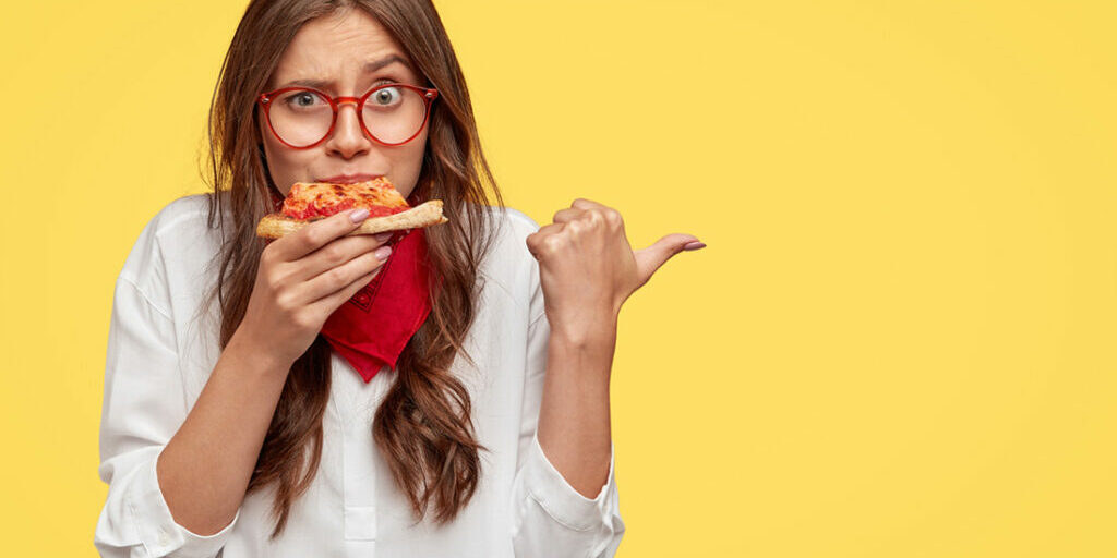 horizontal-shot-surprised-female-eats-tasty-slice-pizza-dressed-fashionable-clothes-indicates-with-thumb-invites-you-pizzeria-isolated-yellow-wall-people-nutrition